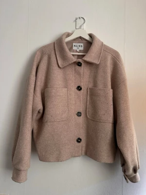 Beige overshirt från NA-KD - Säljer en beige overshirt från NA-KD i storlek 34. Jackan har stora framfickor, klassisk krage och knäpps med fyra mörka knappar. Materialet är mjukt och ull-liknande, perfekt för lager på lager. Overshirten har en avslappnad passform och trendig look.