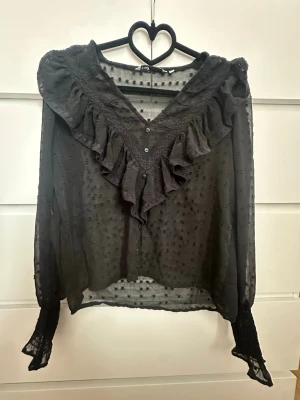 Svart blus med volang och spets - Trendig svart blus från Only med transparent tyg, prickigt mönster och stora volanger längs V-ringningen. Långa ärmar med spetsdetaljer och knappar framtill. 