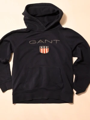 Mörkblå hoodie från GANT - Mörkblå hoodie från GANT med stor huva och magficka. Framtill finns ett broderat GANT-tryck och en sköld med stjärnor och ränder i rött, vitt och blått. Tillverkad i mjukt bomullsmaterial, perfekt för en avslappnad stil.