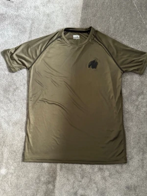 Olivgrön Performance t-shirt - Olivgrön t-shirt från Performance med svart logga på bröstet och tryck på ryggen. Tillverkad i ett lätt och snabbtorkande syntetmaterial, perfekt för aktiva dagar. Klassisk passform med korta ärmar och rund hals.