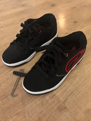 Svarta Heelys sneakers med hjul - Säljer ett par svarta Heelys sneakers med röd logga på sidan och vita sulor. Skorna har inbyggda hjul i sulan och kommer med verktyg för att ta ut hjulen. Perfekta för dig som vill rulla fram med stil! Klassisk snörning och cool design. Nypris 500