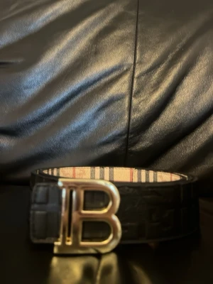Vändbart Burberry bälte med B-spänne - Snyggt vändbart bälte från Burberry med stort B-spänne i metall. Ena sidan är svart med präglat mönster och den andra har klassiskt rutigt Burberry-mönster i beige, rött och svart. Perfekt accessoar för att lyfta din outfit. Den är 115 cm lång