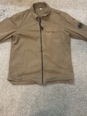  Brun overshirt från C.P. Company - Snygg brun overshirt från C.P. Company med dragkedja framtill, bröstficka och ikonisk svart linsdetalj på ärmen. Tillverkad i lätt bomullsmaterial och har en avslappnad passform. Perfekt för lager på lager och streetwear-stil.