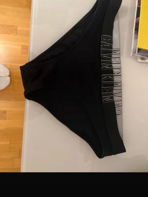 Svart bikiniunderdel Calvin Klein - Snygg svart bikiniunderdel från Calvin Klein med bred resår i midjan där loggan är tryckt i vitt. Klassisk och enkel modell som passar till det mesta. Materialet känns mjukt och stretchigt för en bekväm passform.