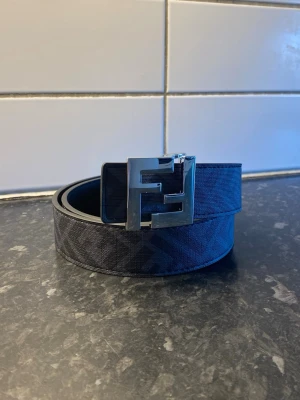 Svart  bälte med silverdetaljer - Snyggt svart bälte  med diskret mönster och stor silvrig FF-logga som spänne. Bältet har en klassisk form och är gjort i skinn med exklusiv känsla. Perfekt accessoar för att lyfta din outfit.