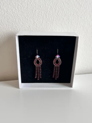 Givenchy örhängen med röda stenar - Säljer ett par äkta statement Givenchy örhängen i silverfärg med röda gnistrande stenar. Örhängena har en droppformad design med fyrkantig detalj upptill och tre hängande kedjor med röda stenar. Perfekta för dig som vill sticka ut och addera färg till din look. Köpte för 3500 kr.