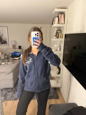 Mörkblå zip hoodie från Peak Performance - Mörkblå hoodie med dragkedja från Peak Performance. Klassisk modell med huva, två fickor framtill och vit logga på bröstet. Tillverkad i mjukt material som passar perfekt för chill dagar. Snygg och enkel att matcha med det mesta.