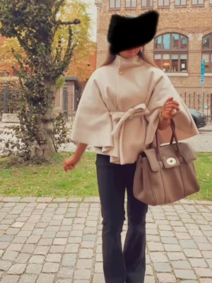 Beige cape-kappa med knytband - Stilren beige cape-kappa med vida ärmar och knytband i midjan. Kappan har hög krage med knappdetalj och en avslappnad, elegant siluett. Perfekt för dig som vill ha en trendig och chic look till hösten.
