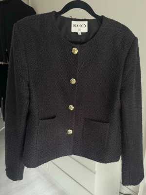 Svart jacka från NAKD - Stilren svart jacka från NAKD med strukturerat tyg och dekorativa guldknappar framtill. Jackan har två fickor och rundad hals. Nypris 799kr.