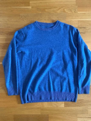 Blå Merinoull tröja med rund hals - En enkel och stilren blå stickad tröja med rund halsringning och ribbade muddar vid ärmslut och nederkant. Perfekt att slänga på sig när det är kyligt ute. Materialet ser mjukt och bekvämt ut, och modellen är klassisk med lång ärm.