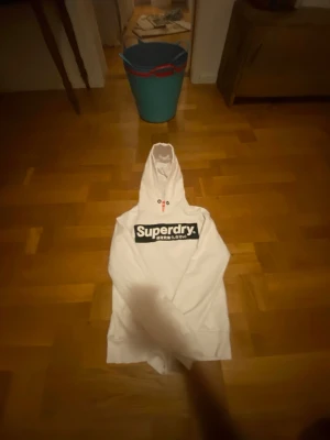 Vit hoodie från Superdry - Snygg vit hoodie från Superdry med stor logga i svart och vitt på bröstet. Klassisk huva och lång ärm, perfekt för en avslappnad streetwear-look. Tillverkad i mjukt material som känns skönt mot huden. 