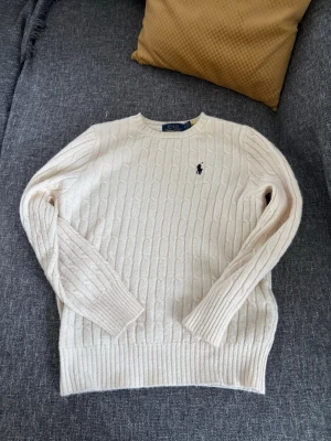 Vit kabelstickad tröja Polo Ralph Lauren - Kabelstickad vit tröja från Polo Ralph Lauren med rund halsringning och klassisk svart logga broderad på bröstet. Tröjan har långa ärmar och ribbade muddar vid ärmslut och nederkant. Perfekt för en stilren och avslappnad look.