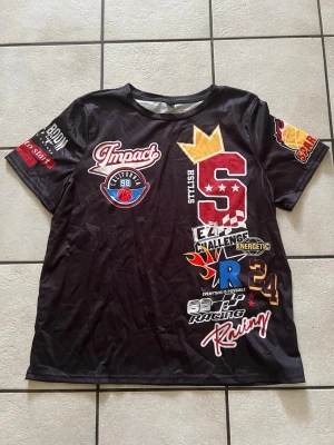 Svart t-shirt med racingtryck storlek XL - Cool svart t-shirt från Impact med massor av färgglada racinginspirerade tryck och loggor på framsidan och ärmarna. T-shirten har korta ärmar och är gjord i ett syntetmaterial som känns lätt och bekvämt. Perfekt för dig som gillar sportiga och unika plagg.