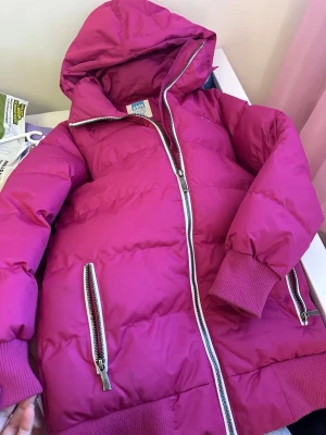 Rosa pufferjacka med huva från WAVE - Snygg rosa pufferjacka från WAVE med stor huva, breda kanaler och silvriga dragkedjor. Jackan har ribbade muddar vid ärmsluten och två framfickor med dragkedja. Perfekt för kalla vinterdagar och riktigt skön att bära.