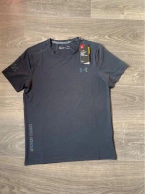 Mörkgrå t-shirt från Under Armour - Snygg mörkgrå t-shirt från Under Armour med diskret logga på bröstet och tryck längs sidan. Klassisk rund hals och korta ärmar. Perfekt för träning eller chill dagar. Tillverkad i mjuk polyester som andas. Pris kan diskuteras 