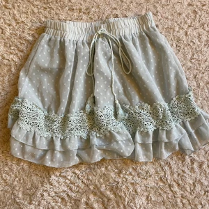 Ljusblå prickig kjol med spets - Supersöt ljusblå kortkjol med vita prickar, volangkant och spetsdetaljer. Kjolen har resår i midjan och knytband framtill för extra passform.