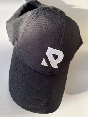 Svart keps från Racqet - Snygg svart keps från Racqet med en vit broderad logga framtill. Klassisk böjd skärm och justerbar baksida. Tillverkad i ett slitstarkt tyg med diskret struktur. Perfekt för en sportig och avslappnad stil.