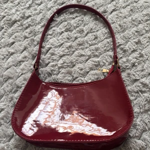 Vinröd axelväska i lack - Snygg vinröd handväska i glansigt lackmaterial med enkel axelrem och guldfärgade metalldetaljer. Väskan har en mjukt rundad form och stängs med dragkedja upptill. Perfekt för dig som vill ha en trendig accessoar med retrovibbar.