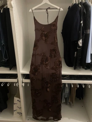 Summer Ready Maxi Dress - White Fox Boutique  - Säljer en elegant brun långklänning med tunna axelband och öppen rygg. Klänningen har ett vackert blommigt mönster i samma ton och är tillverkad i ett glansigt material som ger en lyxig känsla. Klänning är använd enbart en gång, det finns inga skador och den är insydd till storlek M.