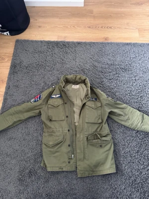 Olivgrön vindjacka med patches - Säljer en olivgrön vindjacka från Ralph Lauren med flera coola patches och broderade detaljer på ärmar och bröst. Jackan har fyra fickor framtill, dragkedja och knappar samt hög krage. Perfekt för dig som gillar militärinspirerad stil.