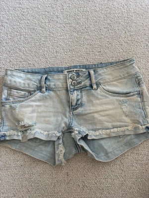 Ljusblå jeansshorts från Tally Weijl - Säljer ett par ljusblå jeansshorts från Tally Weijl med fransiga kanter och slitna detaljer. Shortsen har fem fickor, bälteshällor och stängs med dubbla knappar och dragkedja. Perfekta för sommaren och ger en avslappnad vibe.