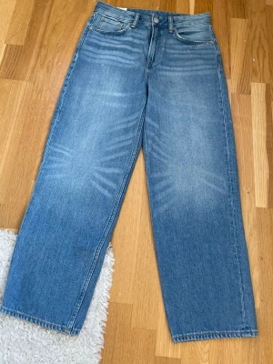 Blå raka jeans med hög midja - Säljer ett par klassiska blå jeans med rak passform och hög midja. Jeansen har fem fickor, knappgylf och snygga slitningar framtill för en avslappnad look. Tillverkade i denim med en ljus tvätt. Perfekta till en trendig vardagsstil.