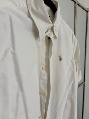 Vit skjorta från Ralph Lauren XS - Klassisk vit kortärmad  skjorta från Ralph Lauren med broderad logga på bröstet. Skjortan har knappar framtill och en snygg krage. Tillverkad i mjuk bomull och passar perfekt till många olika stilar.