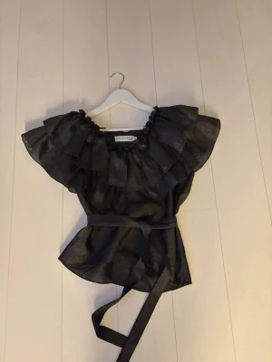 Svart volangblus från Ida Sjöstedt - Supersnygg svart blus från Ida Sjöstedt med stora volanger runt halsen och ett knytband i midjan. 