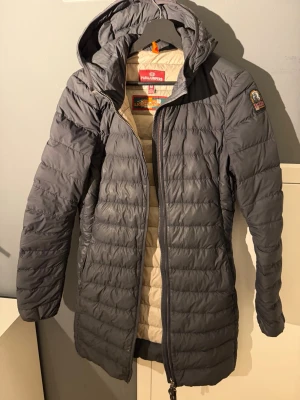 Grå dunjacka med huva från Parajumpers - Snygg grå dunjacka från Parajumpers med huva och quiltad design. Jackan har dragkedja framtill, patch på ärmen och ljus insida. Perfekt för kalla vinterdagar och riktigt skön att bära. Klassisk och stilren modell som funkar till det mesta.