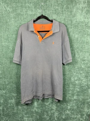 Grå pikétröja från Polo Ralph Lauren - Säljer en grå pikétröja från Polo Ralph Lauren med orange insida på kragen och knappslå. Klassisk pikékrage och korta ärmar. Liten broderad logga på bröstet. Perfekt för en avslappnad men ändå stilren look.