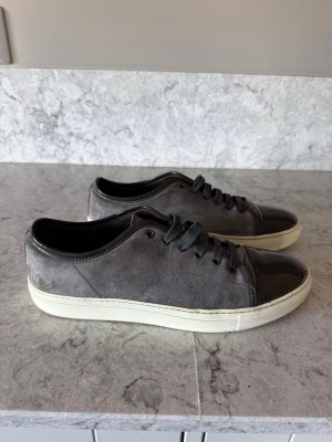 Lanvin Captoe  - Snygga sneakers i grå mocka med svart, glansig tå och svarta skosnören. Skorna har en vit sula och en stilren, låg profil som passar till många olika outfits. Perfekt för dig som gillar en clean och modern look.
