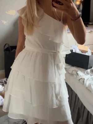 Vit oneshoulder klänning med volanger - Säljer en vit oneshoulder klänning med volanger och knyt i midjan. Klänningen har ett lager-på-lager utseende och är tillverkad i ett lätt, flödande material som passar perfekt till sommaren.