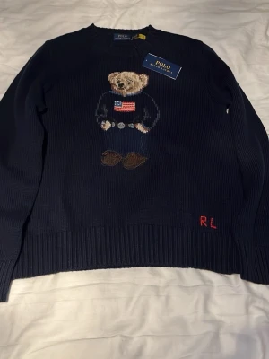 Mörkblå stickad tröja Polo Bear - Mörkblå stickad tröja från Polo Ralph Lauren med den ikoniska Polo Bear-björnen broderad på bröstet. Tröjan har rund halsringning, ribbade muddar och RL i rött nedtill. 