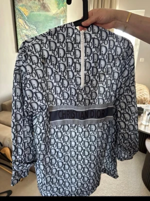 vindjacka  - Snygg vindjacka med ikoniskt blått och vitt monogramtryck över hela plagget. Jackan har halv dragkedja framtill, huva med snören och ett brett. Tillverkad i ett lätt, glansigt material som ger en exklusiv känsla. Passar strl S/M