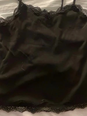 Svart spetslinnetopp - Snygg svart linnetopp med tunna axelband och dekorativ spets längs urringning och nederkant. Perfekt att styla lager på lager eller bära som den är för en chic look.