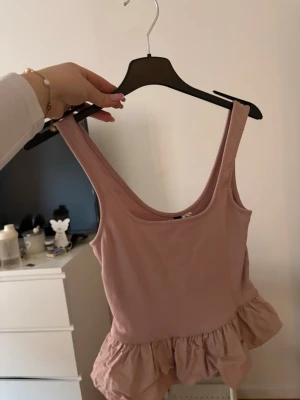 Rosa topp med volangkant - Supersöt rosa topp med breda axelband och djup urringning både fram och bak. Volangkant nedtill ger en lekfull och trendig look. Perfekt att styla med jeans eller kjol för en cool vibe. Mjuk och stretchig bomullskänsla.