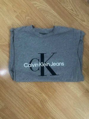 Grå Calvin Klein Jeans t-shirt - Snygg grå t-shirt från Calvin Klein Jeans med stort svart CK-tryck och vit logga på bröstet. Klassisk rund hals och korta ärmar. Tillverkad i mjuk bomull för en skön känsla och enkel stil.