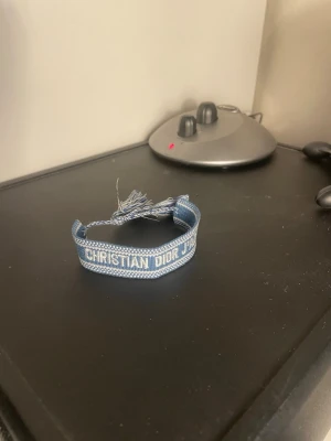 Blått Christian Dior tygarmband - Snyggt blått tygarmband från Christian Dior med texten 'Christian Dior J'Adior' i vitt. Armbandet har justerbar knytning med fransar i ändarna och en stilren, ungdomlig design som passar perfekt till din outfit.