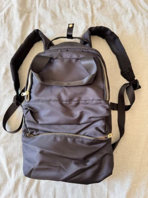 Svart ryggsäck med gulddragkedjor - Snygg svart ryggsäck med guldfärgade dragkedjor och flera fack. Justerbara axelband med mesh för extra komfort och ett praktiskt topphandtag. Nätt modell.