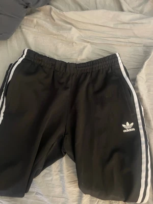 Svarta Adidas mjukisbyxor med vita ränder - Säljer ett par svarta mjukisbyxor från Adidas med klassiska vita ränder längs sidorna och Adidas-logga på vänster ben. Byxorna har resår i midjan och är tillverkade i ett mjukt material som är perfekt för chill dagar.