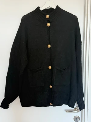 Svart kofta med guldknappar - Missguided, strl M - Stilren svart kofta med ribbad hög krage och sex dekorativa guldknappar framtill. Två framfickor och ribbade muddar vid ärmslut och nederkant. Perfekt för lager på lager och enkel att matcha med olika outfits. 