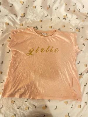 Ljusrosa t-shirt med glittertext barn st 122/128 - Söt ljusrosa t-shirt från Sweet Heart med glittrig guldfärgad text 'girlie' på bröstet. Klassisk rund halsringning och korta ärmar. Perfekt för dig som gillar en enkel men ändå trendig look.