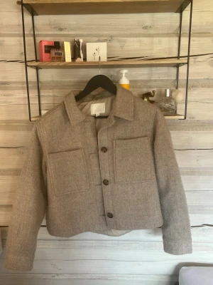 Beige kappa i ull med stora fickor - Snygg beige kappa i ull med stora bröstfickor och knappar framtill. Jackan har en croppad passform, klassisk krage och långa ärmar. 