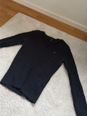 Mörkblå kabelstickad tröja Gant - Kabelstickad mörkblå tröja från Gant med V-ringning och diskret logga broderad på bröstet. Tröjan har långa ärmar och ribbade muddar vid ärmslut och nederkant. Perfekt för en klassisk och stilren look.