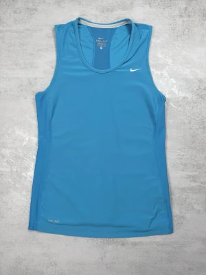 Blått träningslinne från Nike - Snyggt blått träningslinne från Nike med Dri-FIT-material som andas. Linnet har en rund halsringning och meshpaneler på sidorna och ryggen för extra ventilation. Perfekt för gymmet eller löprundan. Liten vit Nike-logga på bröstet.
