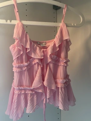Rosa volangtop med knytband - Supersöt rosa topp med tunna axelband och volanger framtill. Toppen har plisserat tyg och knytband i mitten för extra detalj. Perfekt för dig som gillar lite uppklätt och fint