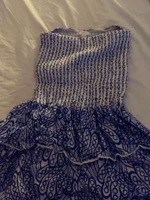 Vit och blå bandeauklänning Gina Tricot - Säljer en söt bandeauklänning från Gina Tricot i vitt och blått. Klänningen har smockad överdel och volangig kjol med ett unikt mönster. Perfekt för varma dagar och sommarens alla äventyr!