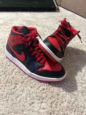 Air Jordan 1 Mid Svart/Röd - Snygga Air Jordan 1 Mid sneakers i svart och rött läder med klassisk röd swoosh och röda skosnören. Skorna har en vit mellansula och ikoniska Jordan-loggor på sidan och plösen. Perfekta för dig som vill sticka ut med en tidlös streetwear-look.