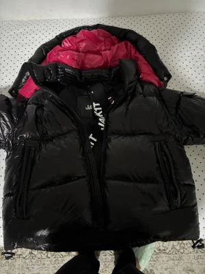Svart dunjacka från JAKT med huva - Snygg svart dunjacka från JAKT med glansig finish och kontrasterande mörkrosa foder. Jag köpte jackan för 2500 nypris och säljer den för detta priset. Vid snabbköp var gärna säkra och prisförslag är alltid välkomna 