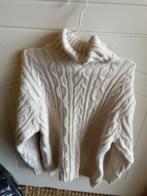 Vit kabelstickad polotröja Rocky - Säljer en chunky vit kabelstickad polotröja från Rocky. Tröjan har hög krage, breda ärmar och ett klassiskt flätmönster över hela plagget. Perfekt för kalla dagar och riktigt mysig att bära. Materialet känns mjukt och tjockt.